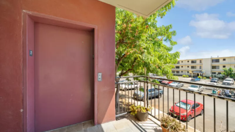 Red elevator door on a balcony with potted plants, overlooking a parking lot, trees, and apartment buildings under a blue sky.