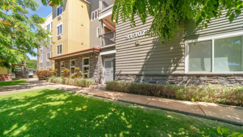 Apartment building with yellow and green siding, stone base, and a lush green lawn under sunny skies.