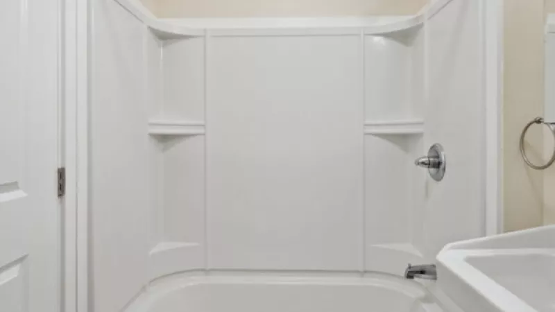Clean white bathtub and shower with chrome fixtures and white wall panels. A white sink is partially visible.