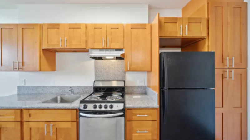 Empty kitchen with light brown wooden cabinets, grey speckled countertops, a stainless steel stove, and a black refrigerator.