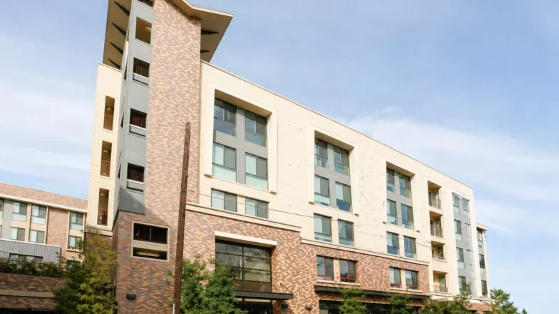 Modern multi-story building with cream, grey, and brick facades, set against a clear sky with some greenery.