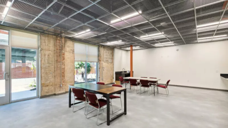 Spacious, modern industrial room with exposed brick walls, polished concrete floor, grid ceiling, and two tables with red chairs.