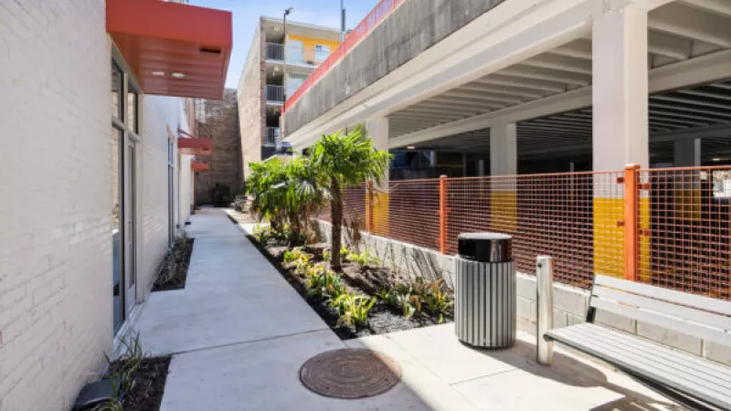 Pathway between white brick building with red awnings and a parking garage with orange fencing. Plants, a bench, and a trash can.