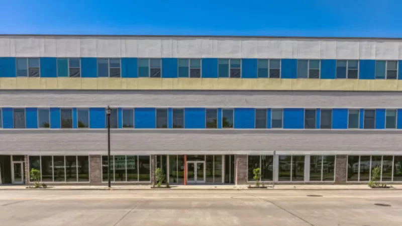 Modern three-story building with grey facade, blue windows, yellow stripe, and ground-level storefronts.