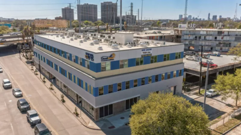 Aerial photograph of a modern three-story building with grey, yellow, and blue accents on a city street corner.