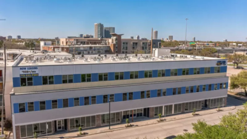 Modern three-story grey and blue commercial building with many windows under a clear sky. City skyline in background.