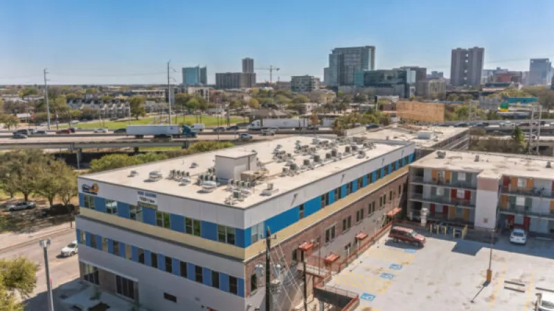Aerial photo of a modern building with blue and yellow trim next to a motel, a highway overpass, and a city skyline.