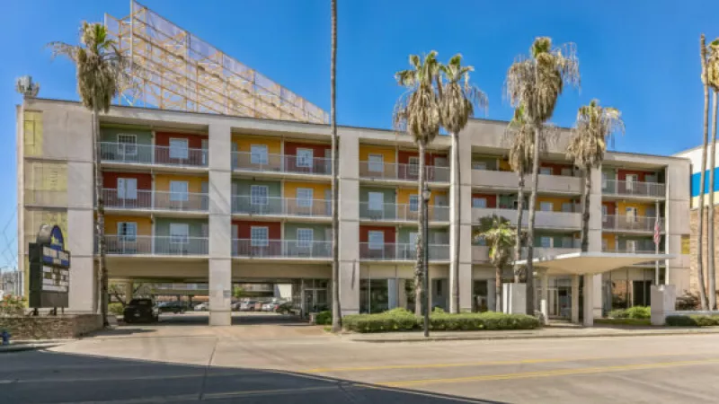 Multi-story apartment building with colorful red, yellow, and green balconies, tall palm trees, and a clear blue sky.