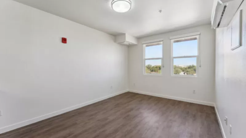 Empty white room with dark wood floor, two windows, overhead light, and a wall-mounted AC unit.
