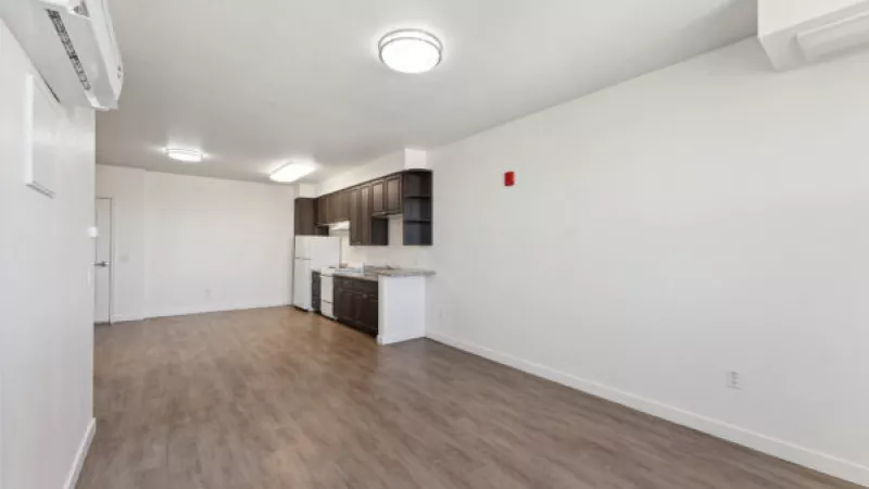 Bright, unfurnished studio apartment with white walls, wood floor, and a compact kitchen with dark cabinets.