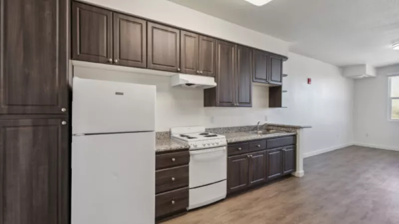 Empty apartment kitchen with dark wood cabinets, white fridge, and stove. Light wood flooring extends into the living area.