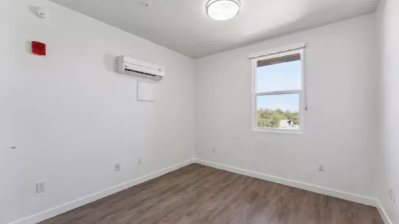 Bright, empty room with white walls, wood-look floor, window, wall-mounted air conditioner, and ceiling light.