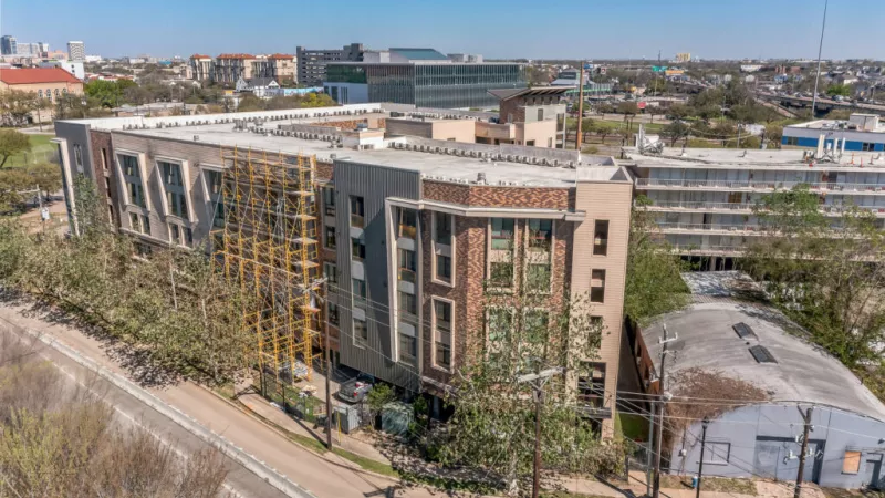Aerial view of a modern multi-story building with varied facades and yellow accents, beside a street with a diverse city background.