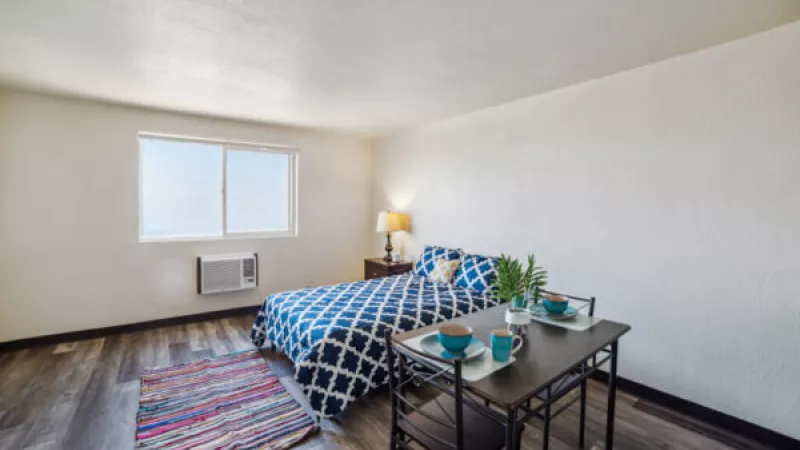 Photograph of a studio apartment with a blue and white patterned bed, small dining table, and colorful rug.