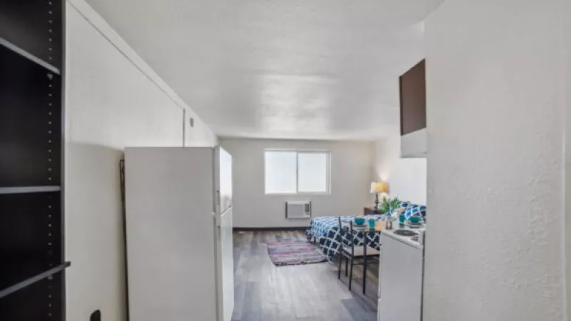 Compact studio apartment with white walls, dark wood floor, bed with checkered cover, refrigerator, and small kitchen by a window.