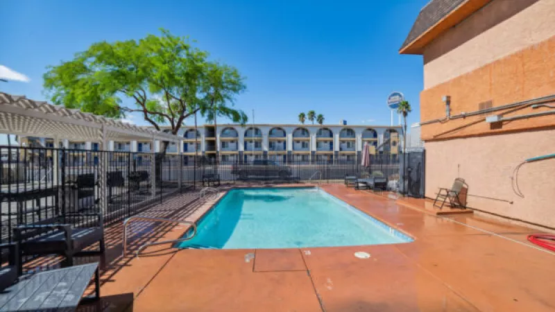 Outdoor swimming pool with blue water, surrounded by a fence, concrete deck, and motel buildings under a sunny sky.