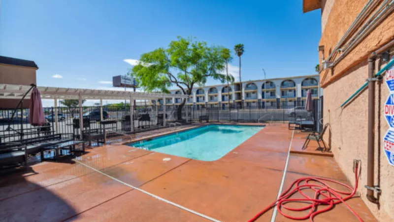 Sunny outdoor motel pool with lounge chairs, red patio, coiled hose, and hotel building in background.