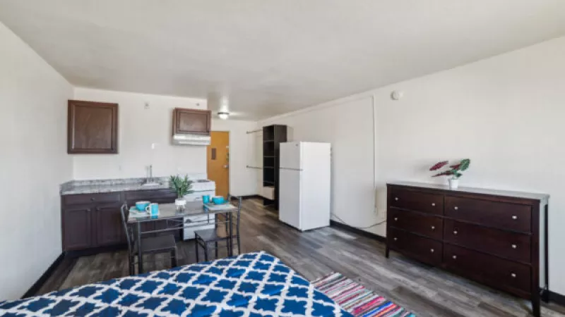 Studio apartment with blue patterned bed, small kitchen, dining table, white fridge, and dark wood dresser.