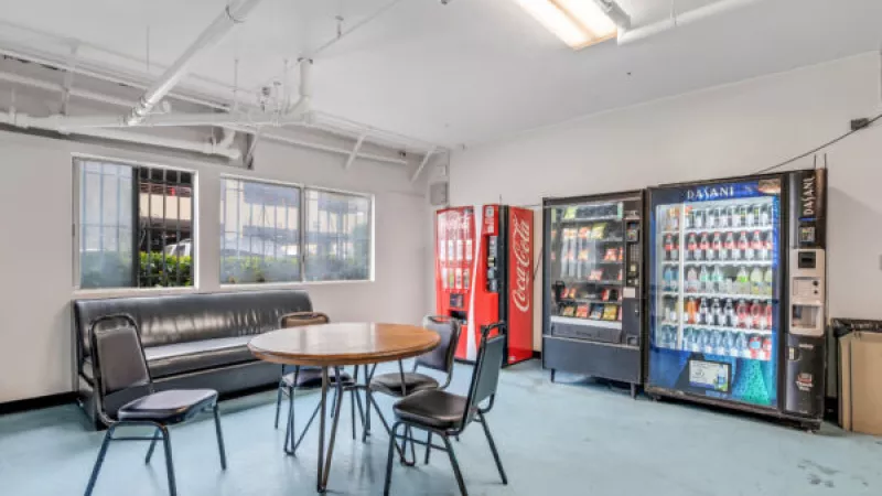 Break room with a black sofa, a round wooden table, three chairs, and three vending machines on a light blue floor.