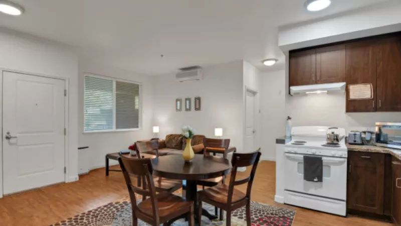 Open-concept apartment living area with a round dining table, a brown sofa, and a kitchen with a white stove.