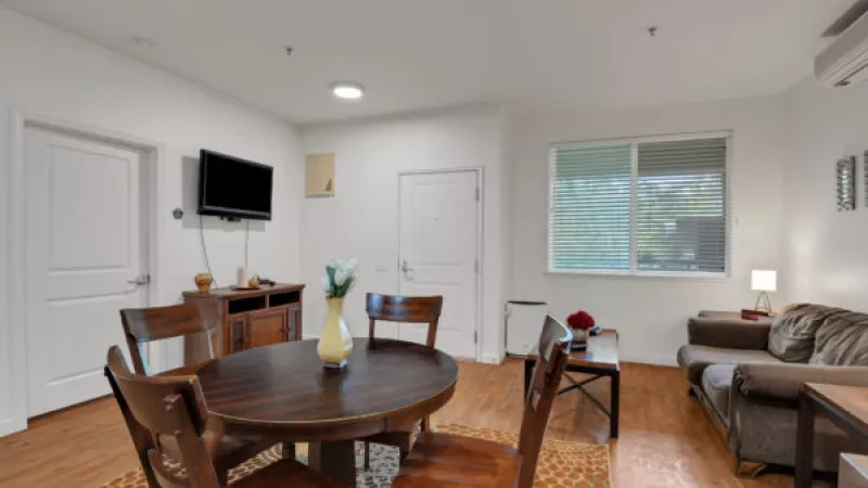 Bright open-plan living and dining area featuring a round wooden table, four chairs, a brown sofa, and a TV on a white wall.