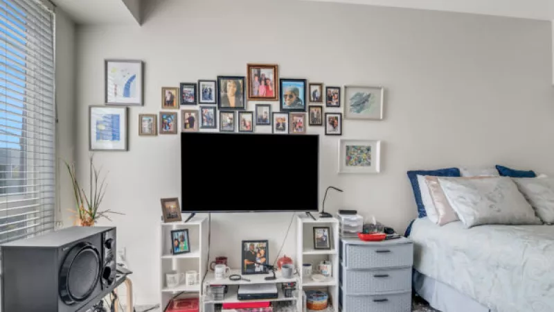 Bedroom with a TV mounted on a wall, surrounded by a large arch of framed photos. A bed with light blue bedding is to the right.