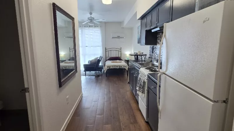 Studio apartment with a galley kitchen on the right, white fridge, and a living area with a bed and couch on the left.