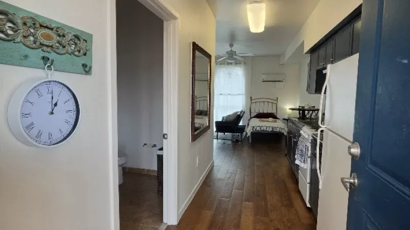 Narrow studio apartment with wood floors. Dark blue kitchen on right, bed at far end. White clock and open doorway on left.