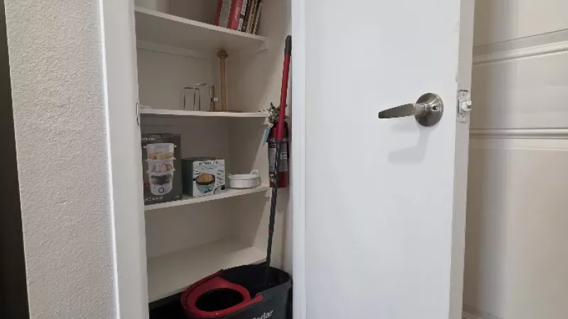Pantry closet with white shelves storing paper towels, cleaning supplies, and a red mop bucket.