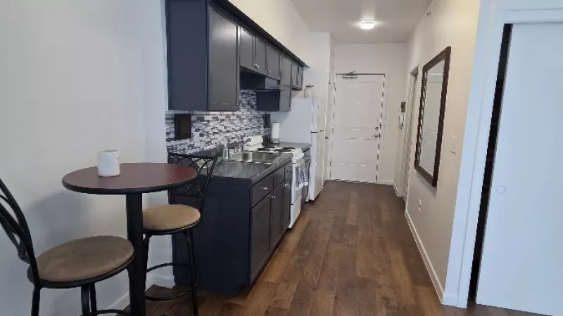Narrow studio apartment interior with a compact kitchen area on the left, small dining table, and dark wood floors extending down a hallway.