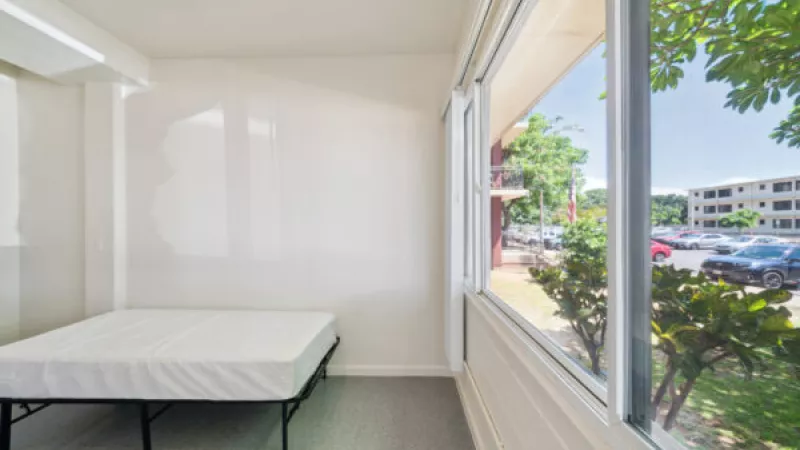 Bright, empty room with a white mattress on a metal bed frame. A large window overlooks a parking lot and buildings.