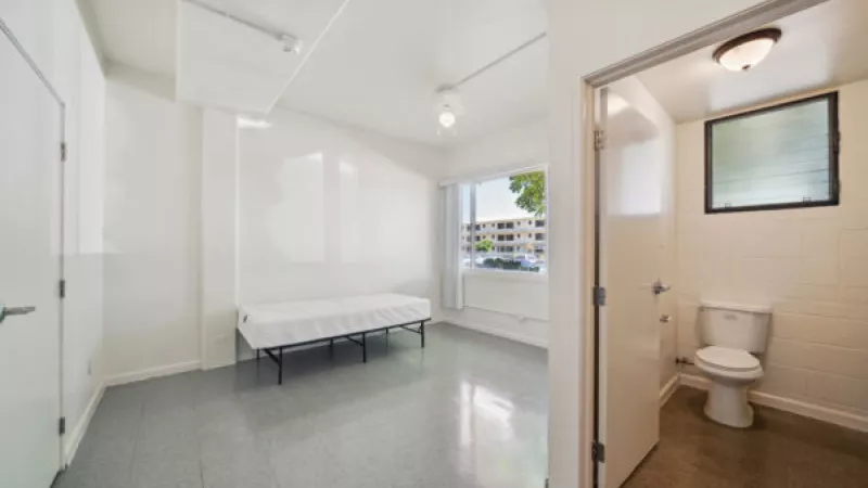 Empty, minimalist room with a single bed, white walls, grey floor, and an open door to a small bathroom with a toilet.