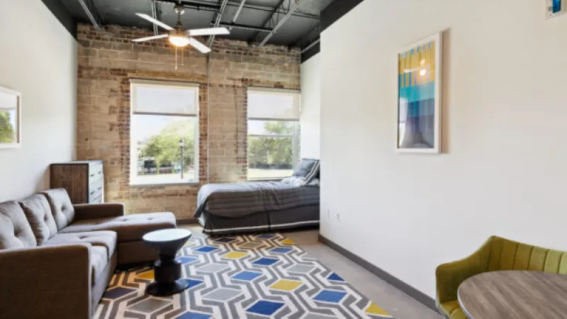 Modern studio apartment with exposed brick wall, brown sectional sofa, grey bed, and a colorful geometric rug.