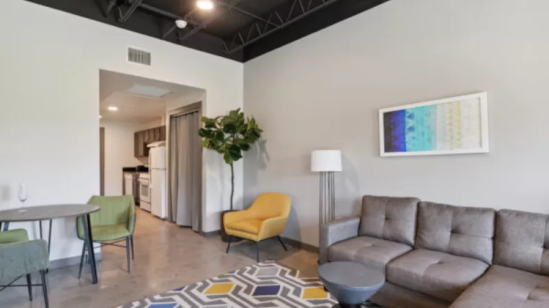 Modern living room with a gray sectional, yellow armchair, colorful geometric rug, and a glimpse of a kitchen.