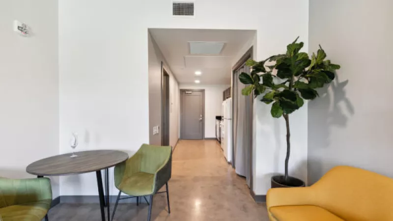Modern entryway with a round table, green chairs, a yellow sofa, and a tall potted plant, leading to a long white hallway.