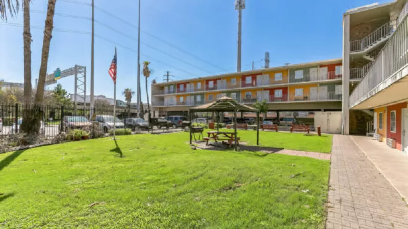 Grassy courtyard with a gazebo and picnic table, adjacent to a colorful two-story motel building under a clear blue sky.