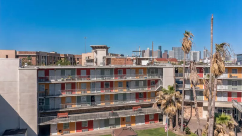 Multi-story apartment building with colorful door facades, palm trees, and a distant city skyline under a blue sky.