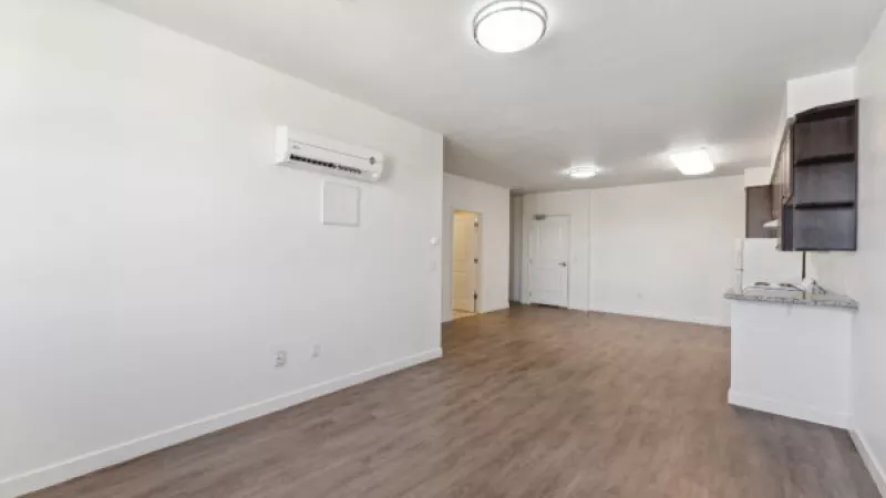 Empty apartment living room with white walls, wood-look floor, a wall-mounted AC unit, and dark kitchen cabinets.