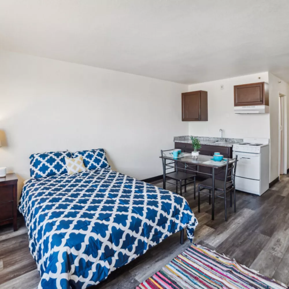 Compact studio apartment with a bed featuring blue and white patterned bedding, a small dining table, and a kitchenette.