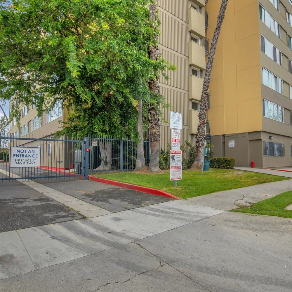 Metal gate blocks a paved entrance to a multi-story building, with green trees and a sidewalk leading past.
