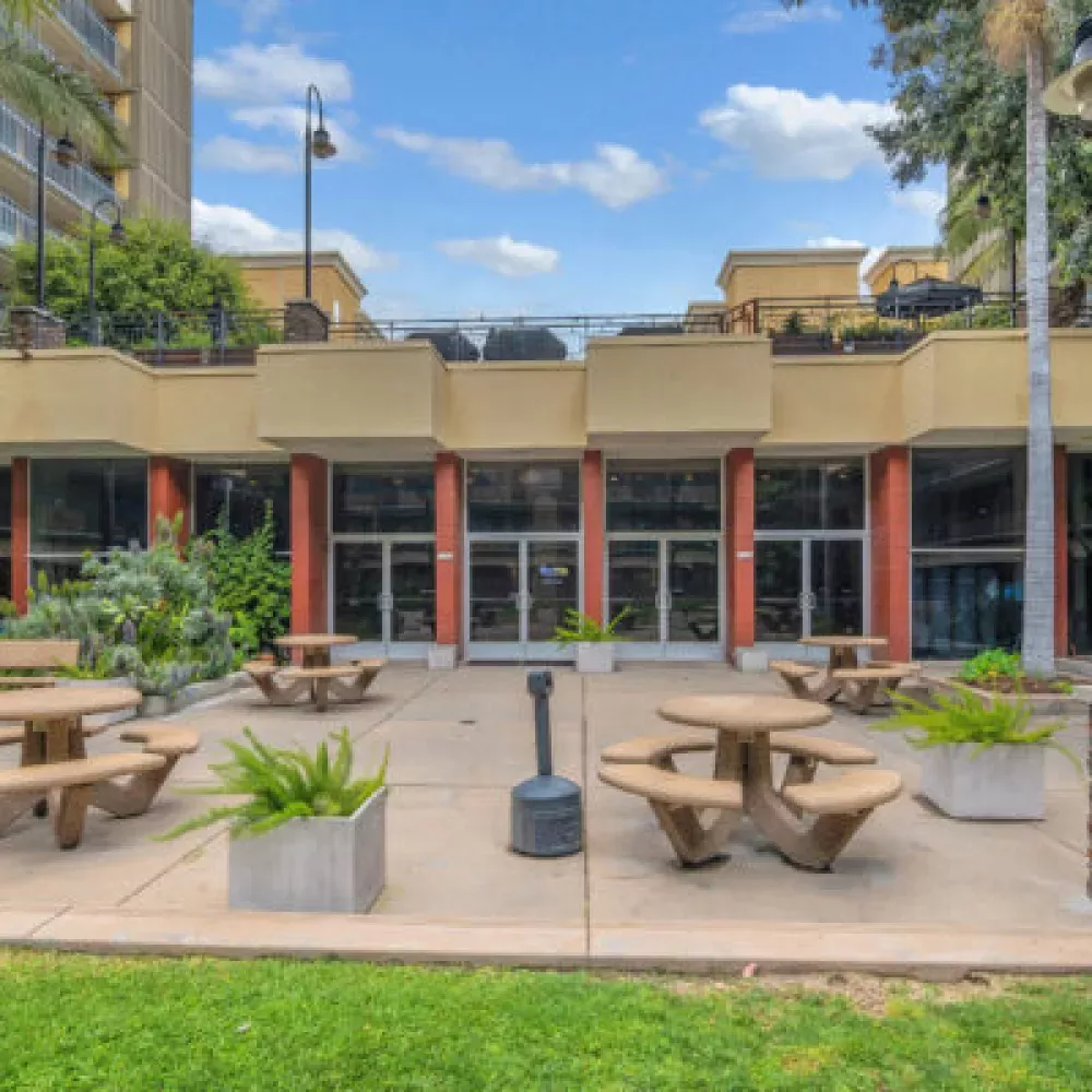 Outdoor courtyard with beige picnic tables, a building with large windows, and residential towers.