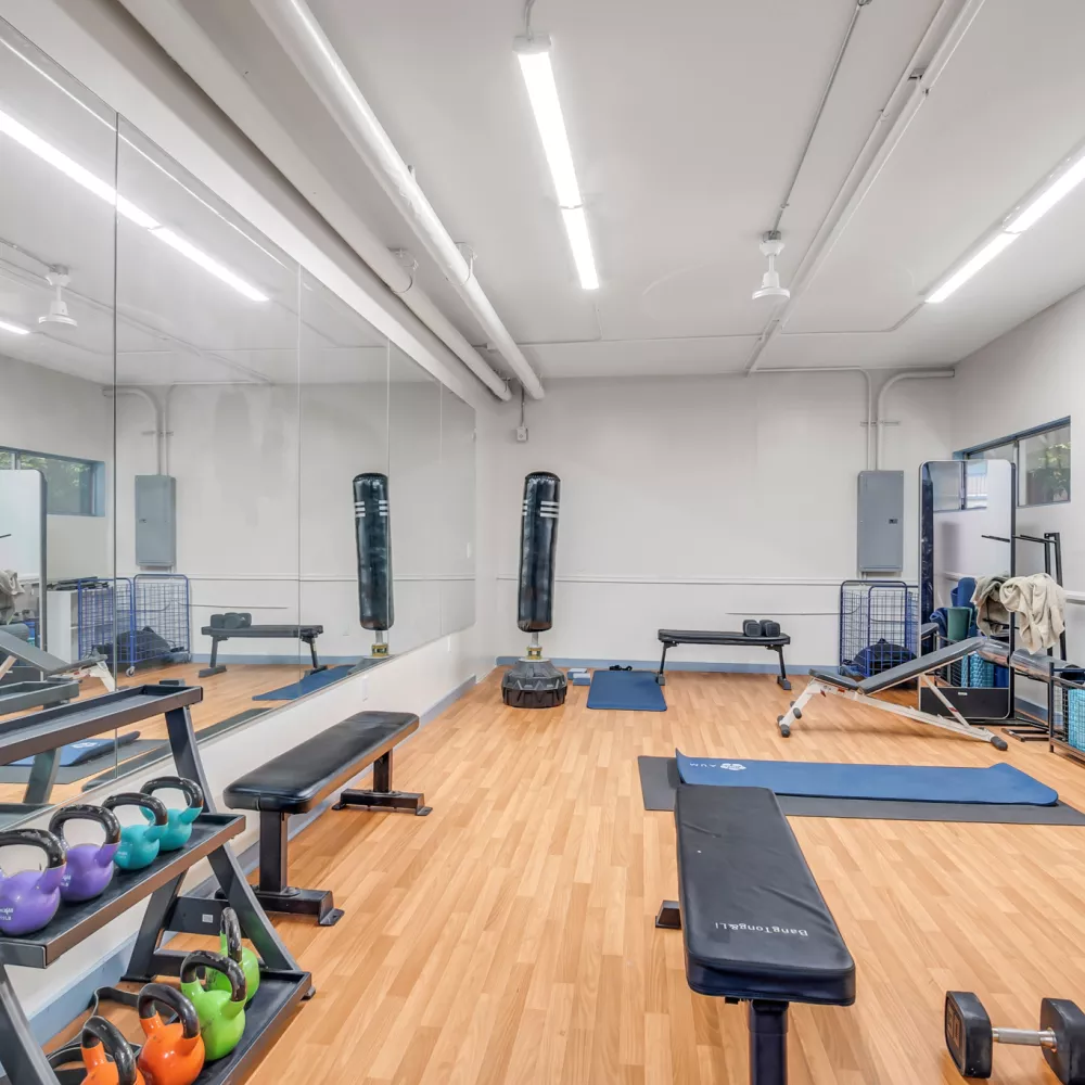 Brightly lit gym with a mirrored wall, wooden floor, weight benches, kettlebells, dumbbells, and exercise mats.