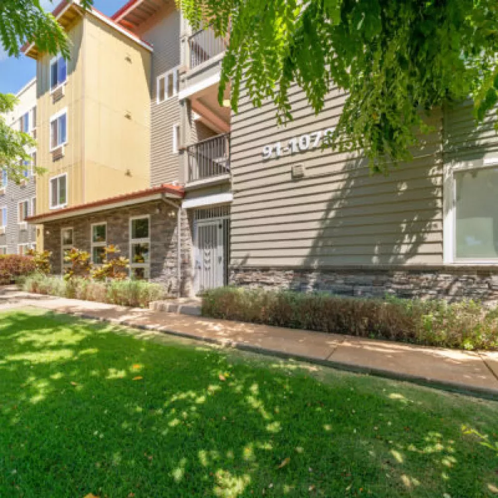Multi-story Kipuka building exterior with yellow and gray siding, stone accents, a green lawn, and sunlit trees.