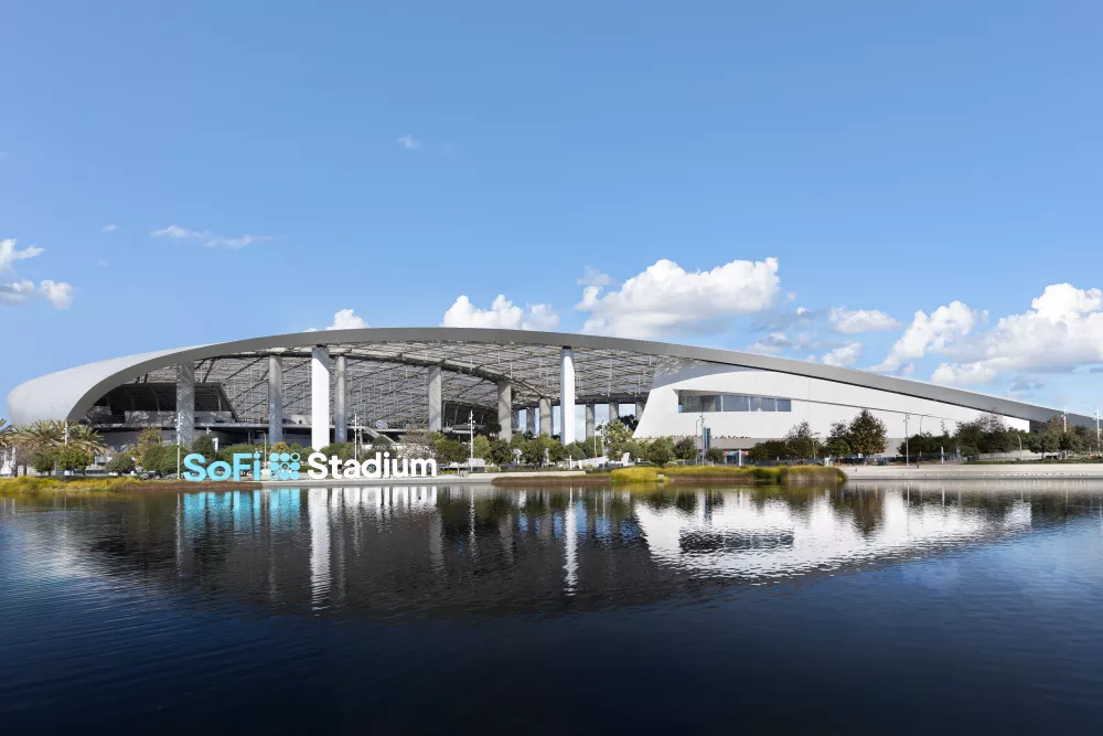 Photograph: SoFi Stadium, a large, modern arena with an arched roof, reflected in dark water under a partly cloudy blue sky.
