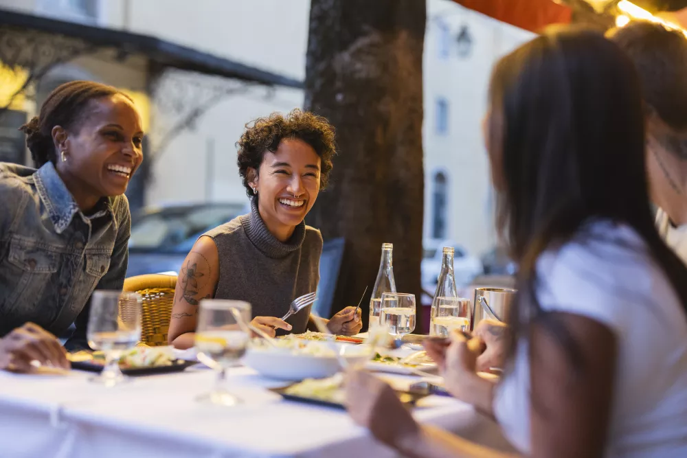 Two women laugh while dining outdoors with friends at a restaurant, lit by evening lights.