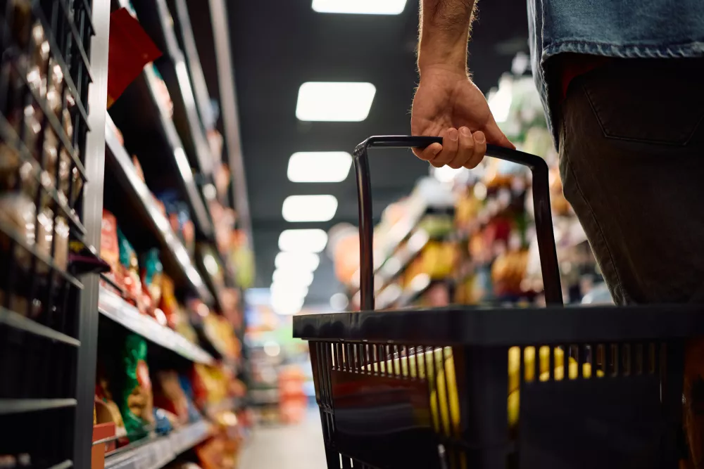 Shopper's hand holds a black basket, walking down a blurred grocery store aisle filled with colorful products.