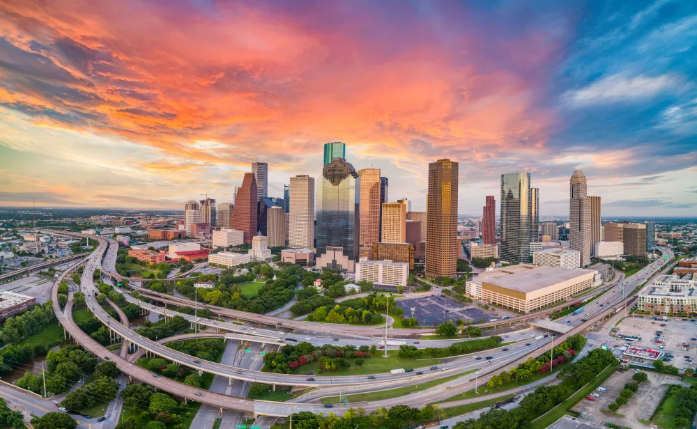 Aerial view of Houston's downtown skyscrapers and a large highway interchange under a fiery sunset sky.