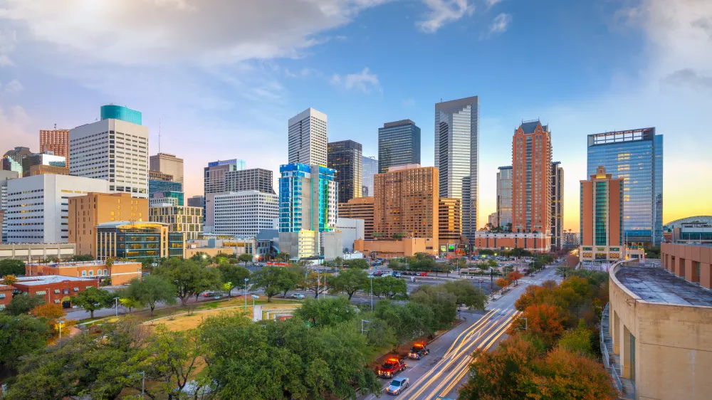 City skyline at dusk, with numerous skyscrapers, green parks, and an elevated road below. The sky shows warm hues.