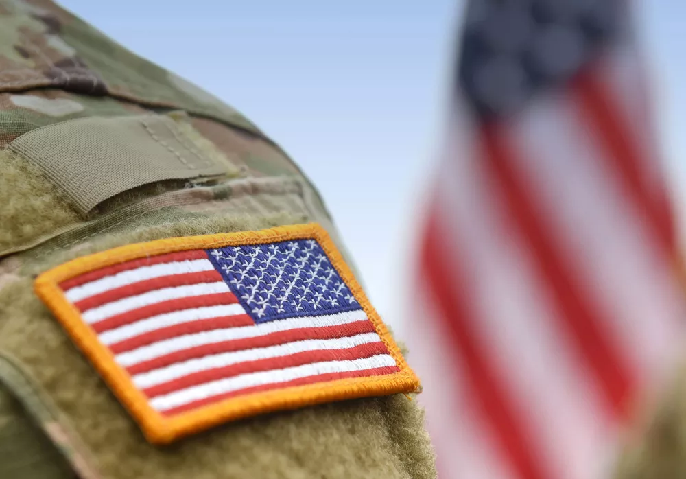 American flag patch on a soldier's camouflage uniform, with a blurred US flag in the background.