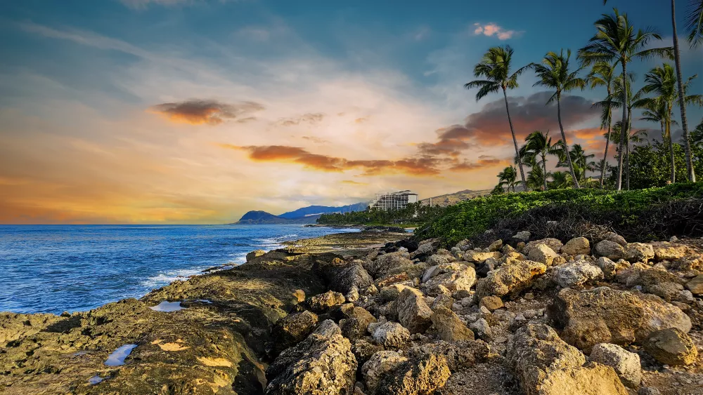 Rocky tropical coastline at sunset. Palm trees and resort buildings line the shore, with waves crashing under a vibrant orange sky.